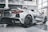 A sleek silver sports car displayed in a modern showroom with large windows. The vehicle features distinctively designed tail lights and black, alloy wheels. The floor of the showroom is polished, reflecting the car's streamlined body.