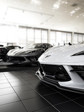 Several sports cars are displayed in a showroom with glossy tiled flooring and a well-lit, modern interior. The cars have sleek, aerodynamic designs and are positioned close to each other.