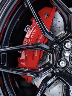 Close-up of a car's disc brake system featuring a vivid red brake caliper and silver perforated rotor. The black glossy wheel rim creates a stark contrast, highlighting the brake components.