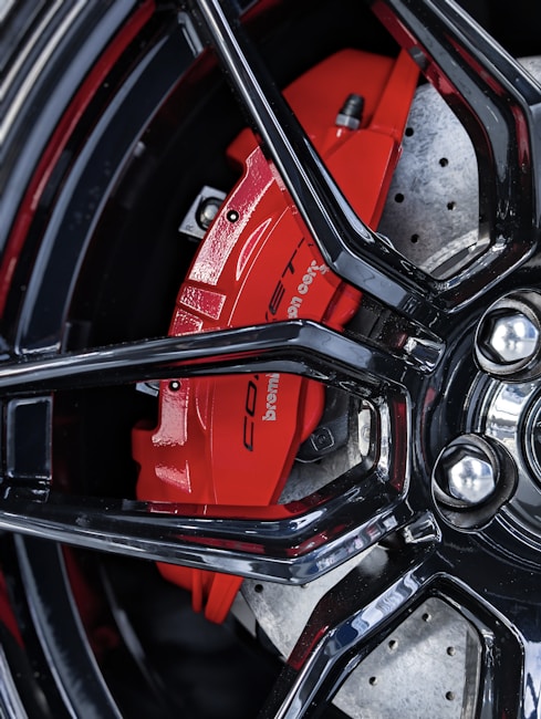 Close-up of a car's disc brake system featuring a vivid red brake caliper and silver perforated rotor. The black glossy wheel rim creates a stark contrast, highlighting the brake components.