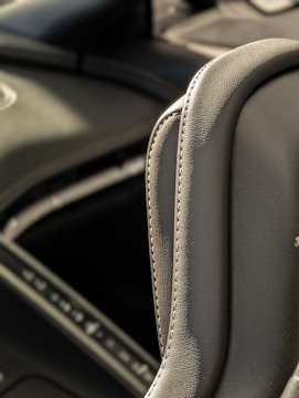 A close-up view of a gray leather car interior, focusing on the stitching detail and texture of the material. The background is slightly blurred, showing parts of the dashboard and console.