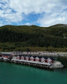 Several red cabins stand on wooden stilts by a serene body of water, with a backdrop of lush green hills and scattered cloud coverage in the sky.