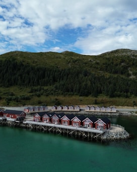 Several red cabins stand on wooden stilts by a serene body of water, with a backdrop of lush green hills and scattered cloud coverage in the sky.