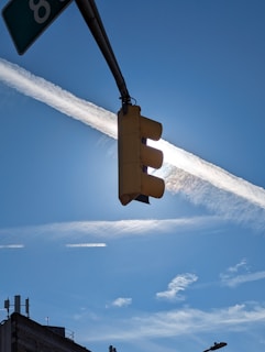 A traffic light is set against a bright blue sky, with a luminous sun partially obscured by a thin, wispy white cloud. The contrail of an airplane cuts across the sky in a straight line, contrasting with the scattered cirrus clouds. The image includes the silhouette of a building topped with various antennas on the lower left.