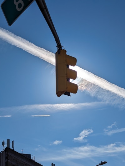 A traffic light is set against a bright blue sky, with a luminous sun partially obscured by a thin, wispy white cloud. The contrail of an airplane cuts across the sky in a straight line, contrasting with the scattered cirrus clouds. The image includes the silhouette of a building topped with various antennas on the lower left.