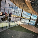 A spacious interior with large, floor-to-ceiling windows allowing ample natural light to fill the space. There is a glossy, reflective floor and several seating arrangements, including rocking chairs. Display stands with merchandise and a small potted plant are visible. The outside view through the windows shows an open landscape under a partly cloudy sky.