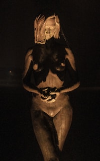 A sculpture of a nude female figure stands in a dim, shadowy environment. The lighting highlights the details of the sculpture, casting dramatic shadows across the form. The figure's posture is neutral, with hands clasped in front of the body and an expressionless face.