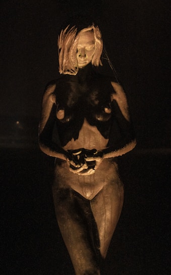 A sculpture of a nude female figure stands in a dim, shadowy environment. The lighting highlights the details of the sculpture, casting dramatic shadows across the form. The figure's posture is neutral, with hands clasped in front of the body and an expressionless face.