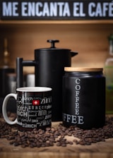 A cozy setup featuring a black coffee cup with 'Zürich' written multiple times in various fonts next to a matte black French press and a similarly styled black coffee container labeled 'COFFEE'. Coffee beans are scattered around them on a wooden surface. The background includes a sign with the phrase 'ME ENCANTA EL CAFE' in bold white letters.