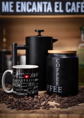 A cozy setup featuring a black coffee cup with 'Zürich' written multiple times in various fonts next to a matte black French press and a similarly styled black coffee container labeled 'COFFEE'. Coffee beans are scattered around them on a wooden surface. The background includes a sign with the phrase 'ME ENCANTA EL CAFE' in bold white letters.