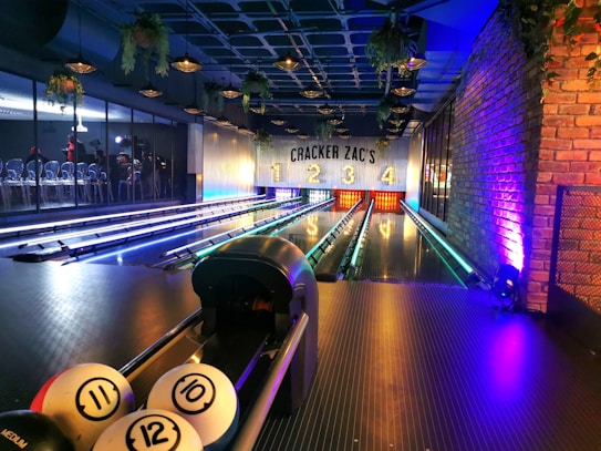 A modern indoor bowling alley with four lanes ending at a wall labeled with 'Cracker Zac's' and lane numbers 1 to 4. The lanes are lit with colorful lights and are bordered by lush green plants hanging from the ceiling. A section on the left shows people seated, possibly waiting their turn to play. The atmosphere is stylish with a combination of contemporary design elements and warm lighting.