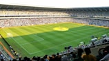 A large soccer stadium filled with a cheering crowd. The field is well-maintained with vibrant green grass, and players can be seen scattered on the pitch. The atmosphere is lively and bustling, typical of a sports event.