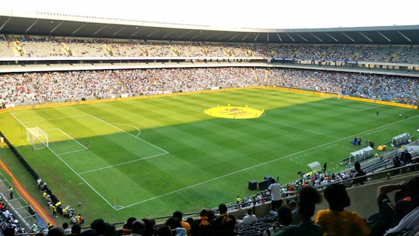 A large soccer stadium filled with a cheering crowd. The field is well-maintained with vibrant green grass, and players can be seen scattered on the pitch. The atmosphere is lively and bustling, typical of a sports event.