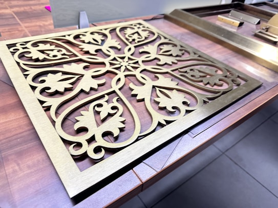 An ornate, gold-colored metal panel features intricate, symmetrical patterns resembling floral and leaf motifs. It is set on a wooden surface with other similar metallic elements nearby.