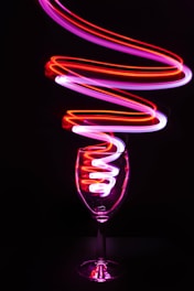 A wine glass is positioned in the center with vibrant light trails in shades of red and pink swirling around and inside it, creating a dynamic and artistic effect against a dark background.