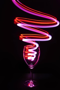 A wine glass is positioned in the center with vibrant light trails in shades of red and pink swirling around and inside it, creating a dynamic and artistic effect against a dark background.