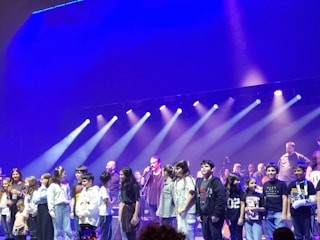 A group of children and adults stand on stage under bright spotlights. The stage is illuminated with purple and blue lighting, creating a vibrant atmosphere.