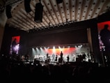 An orchestra is performing on stage with a large, illuminated backdrop. The stage lighting highlights the silhouettes of musicians playing various instruments. The atmosphere is grand, and the ceiling has patterned designs. There are large monitors on either side of the stage showing a closer view of the performers.
