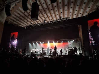 An orchestra is performing on stage with a large, illuminated backdrop. The stage lighting highlights the silhouettes of musicians playing various instruments. The atmosphere is grand, and the ceiling has patterned designs. There are large monitors on either side of the stage showing a closer view of the performers.