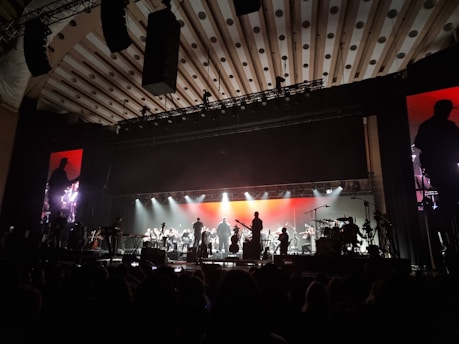 An orchestra is performing on stage with a large, illuminated backdrop. The stage lighting highlights the silhouettes of musicians playing various instruments. The atmosphere is grand, and the ceiling has patterned designs. There are large monitors on either side of the stage showing a closer view of the performers.
