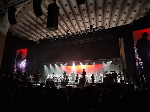 An orchestra is performing on stage with a large, illuminated backdrop. The stage lighting highlights the silhouettes of musicians playing various instruments. The atmosphere is grand, and the ceiling has patterned designs. There are large monitors on either side of the stage showing a closer view of the performers.