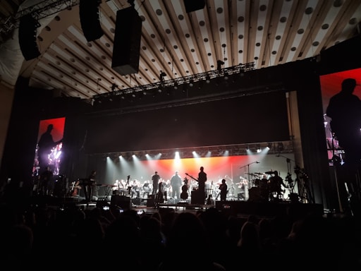An orchestra is performing on stage with a large, illuminated backdrop. The stage lighting highlights the silhouettes of musicians playing various instruments. The atmosphere is grand, and the ceiling has patterned designs. There are large monitors on either side of the stage showing a closer view of the performers.