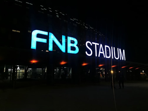Large illuminated signage displays the letters 'FNB STADIUM' in bright colors against a dark background. The letters are prominently lit, creating a vivid contrast with the surrounding evening or night setting.