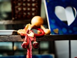 A decorative element with red and gold bells and ribbons is placed on a wooden shelf. In the background, a blue frame with heart-shaped cutouts is visible, creating an artistic and warm atmosphere.