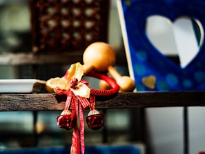 A decorative element with red and gold bells and ribbons is placed on a wooden shelf. In the background, a blue frame with heart-shaped cutouts is visible, creating an artistic and warm atmosphere.