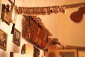 Several violins are hanging on a horizontal rack attached to a wall. The wall is adorned with framed photographs and other musical instruments. The room has a warm, cozy atmosphere highlighted by soft lighting.