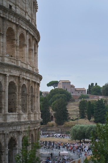 A close-up view of the Colosseum's arches with its ancient stone architecture visible. In the background, there is a green landscape with trees and an old building. A large group of tourists is gathered near the structure, indicating it's a popular historical site.