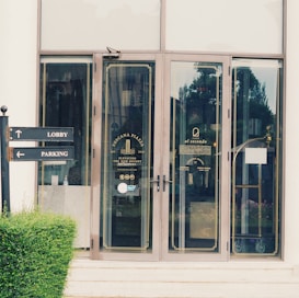 A glass door entrance to a building with signage indicating 'Toscana Piazza' and 'al secondo.' The door features a decorative gold outline and information about the premises. Placed nearby are signs pointing towards 'Lobby' and 'Parking,' set amidst neatly trimmed greenery.