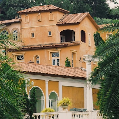 A Mediterranean-style house with warm terracotta walls and multiple levels. The building features arched windows and doors, as well as a red-tiled roof. Lush greenery and palm trees surround the property, adding to the serene and tropical ambiance.