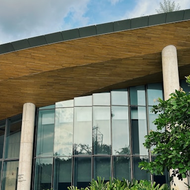 A modern building featuring a sleek design with large glass windows and a wooden overhang. The structure includes tall cylindrical concrete pillars. Green foliage is visible in the foreground, providing a natural contrast to the architectural elements.