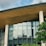 A modern building featuring a sleek design with large glass windows and a wooden overhang. The structure includes tall cylindrical concrete pillars. Green foliage is visible in the foreground, providing a natural contrast to the architectural elements.