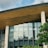 A modern building featuring a sleek design with large glass windows and a wooden overhang. The structure includes tall cylindrical concrete pillars. Green foliage is visible in the foreground, providing a natural contrast to the architectural elements.