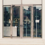 Glass doors with gold trim leading into a cafe, featuring branding and information for Toscana Piazza and Al Secondo. Reflections of the surroundings and some interior lights are visible on the glass.