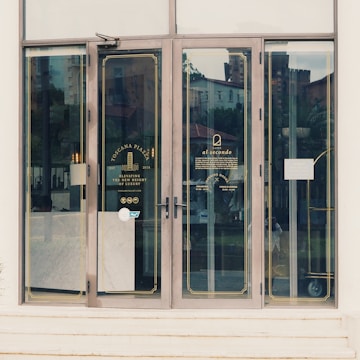 Glass doors with gold trim leading into a cafe, featuring branding and information for Toscana Piazza and Al Secondo. Reflections of the surroundings and some interior lights are visible on the glass.