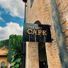A black wooden sign with golden letters spelling 'Primo Cafe' is mounted on a brick wall. The building has a rustic appearance with aged brickwork. In the background, there are leafy green plants and a clear blue sky with some clouds.