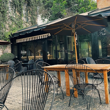 A charming outdoor cafe setting with iron chairs and wooden tables on a cobblestone patio. Large umbrellas provide shade, and the background features a building with ivy-covered walls and window awnings.