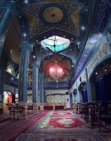 A richly decorated interior featuring intricate geometric patterns and colorful tile work on the walls and ceiling. A grand chandelier hangs prominently, with ornate rugs covering the floor. The ambience is serene, with a combination of blue, yellow, and red tones dominating the space.