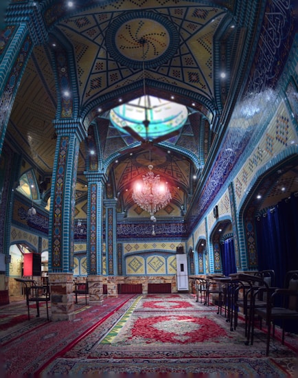A richly decorated interior featuring intricate geometric patterns and colorful tile work on the walls and ceiling. A grand chandelier hangs prominently, with ornate rugs covering the floor. The ambience is serene, with a combination of blue, yellow, and red tones dominating the space.