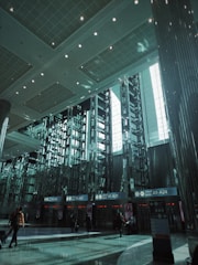 A spacious modern airport terminal features high ceilings and geometric designs. Rows of tall metallic structures and reflective surfaces dominate the scene, while natural light filters through large windows. People with luggage move toward the gates labeled A1-A24.