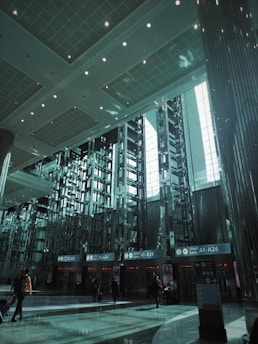 A spacious modern airport terminal features high ceilings and geometric designs. Rows of tall metallic structures and reflective surfaces dominate the scene, while natural light filters through large windows. People with luggage move toward the gates labeled A1-A24.