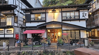 A traditional Japanese building with a distinctive architectural style, featuring wooden lattice windows and a prominent red awning. The entrance is warmly lit, with a variety of plants and small trees arranged in front. The structure is surrounded by other similar buildings, suggesting a quaint, historic district.