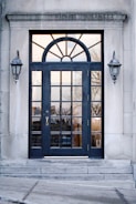 A black-framed double door with multiple glass panels is set in a stone building. Above the door is an arched window with sunburst-style divisions. On either side of the door, there are two classic wall-mounted lanterns. The reflection in the glass panels shows an outdoor scene with trees and buildings.