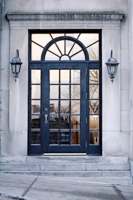 A black-framed double door with multiple glass panels is set in a stone building. Above the door is an arched window with sunburst-style divisions. On either side of the door, there are two classic wall-mounted lanterns. The reflection in the glass panels shows an outdoor scene with trees and buildings.