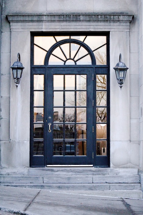 A black-framed double door with multiple glass panels is set in a stone building. Above the door is an arched window with sunburst-style divisions. On either side of the door, there are two classic wall-mounted lanterns. The reflection in the glass panels shows an outdoor scene with trees and buildings.