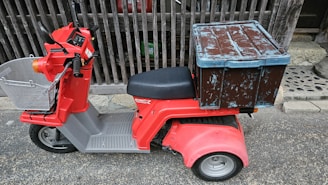 A red three-wheeled scooter with a large, weathered blue and brown storage box attached to the back. The scooter has a basket on the front, handlebars, and a cushioned seat. It is parked on a textured street surface, adjacent to a wooden fence and stone pavement.