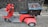 A red three-wheeled scooter with a large, weathered blue and brown storage box attached to the back. The scooter has a basket on the front, handlebars, and a cushioned seat. It is parked on a textured street surface, adjacent to a wooden fence and stone pavement.